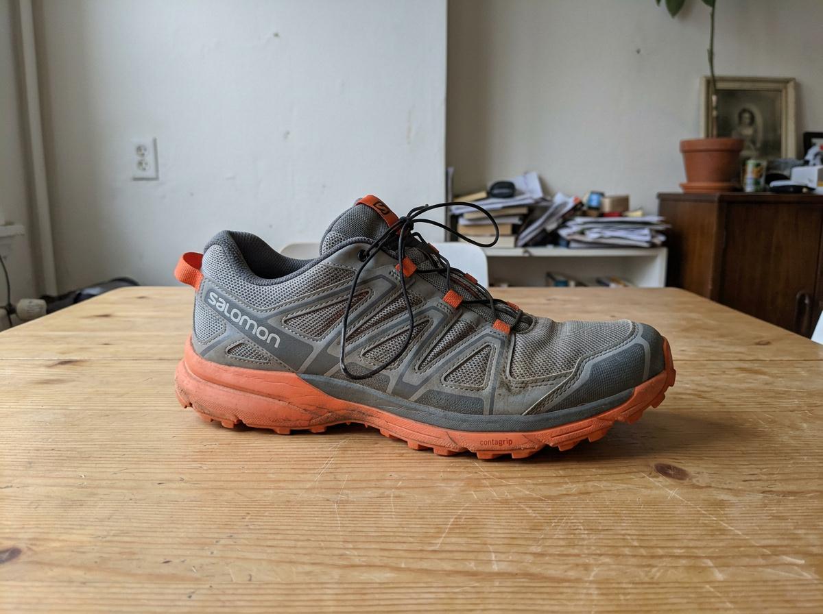 A plain phone photo of a grey and orange trail running shoe on a wooden table